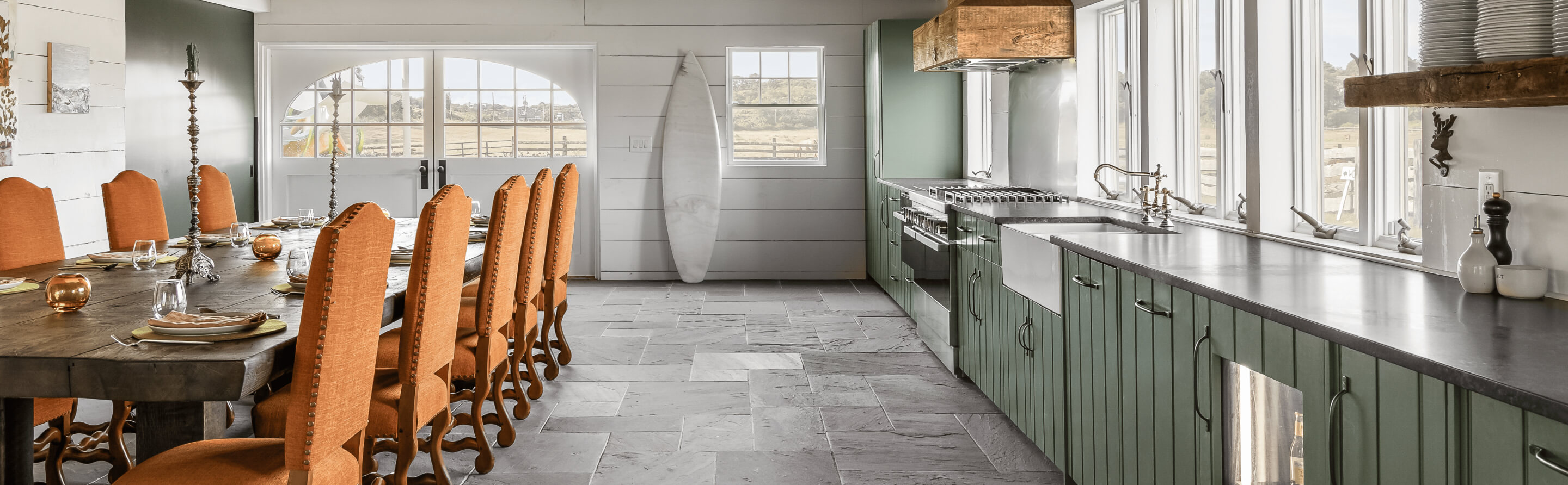 Open kitchen with green cabinetry, stone flooring, custom wood vent hood and a spacious dining table for entertaining.