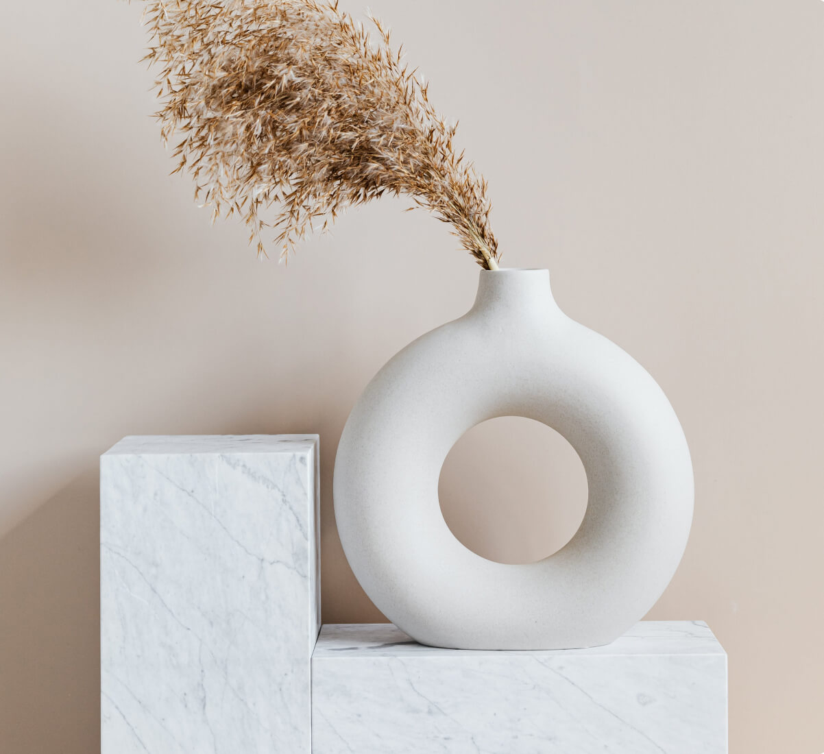A round, donut-like vase sitting on a stone display platform with dried florals in it. A round, donut-like vase sitting on a stone display platform with dried florals in it.