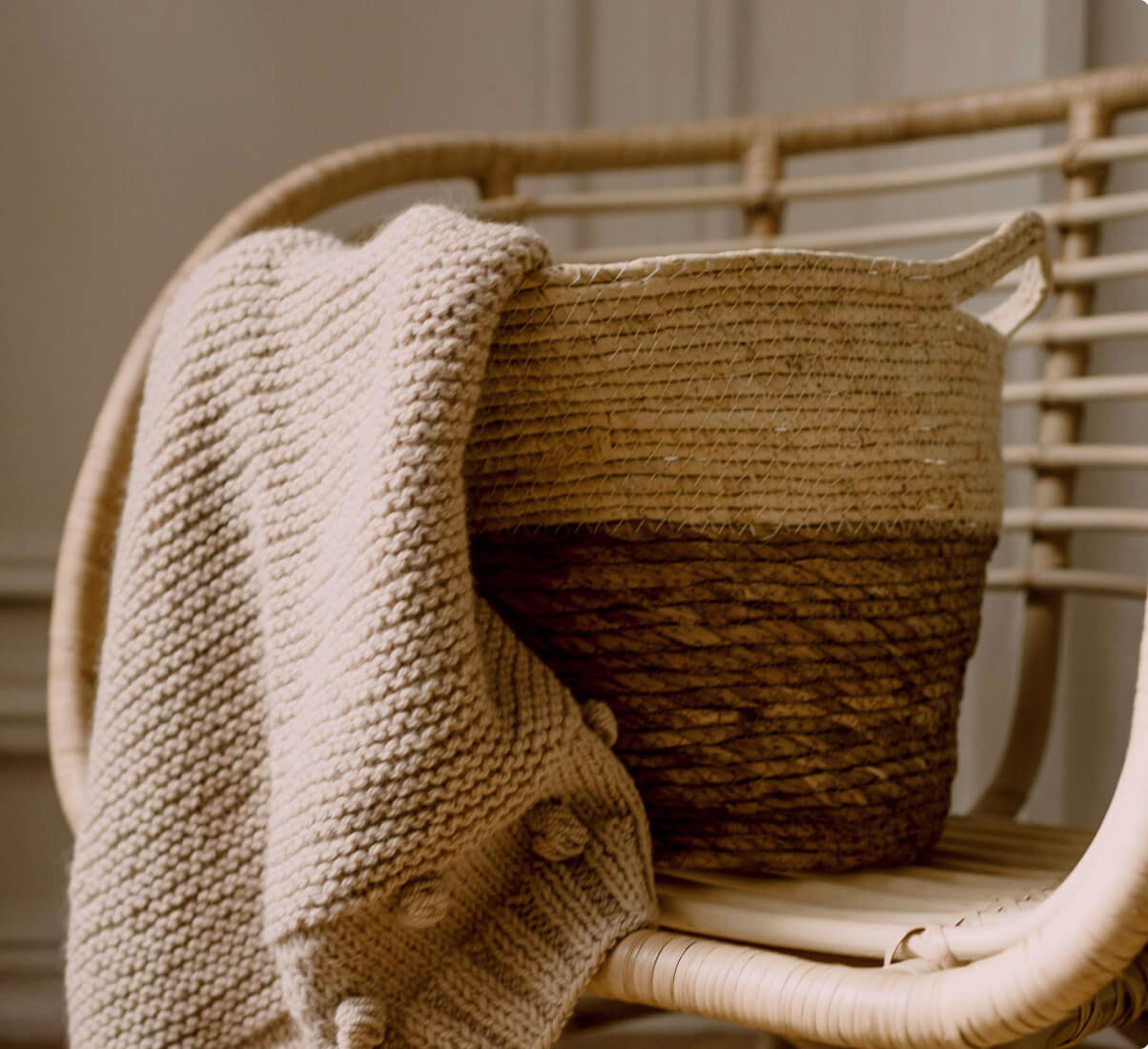 A rattan chair paired with a natural-fiber basket and blanket in varying shades of beige and brown. A rattan chair paired with a natural-fiber basket and blanket in varying shades of beige and brown.